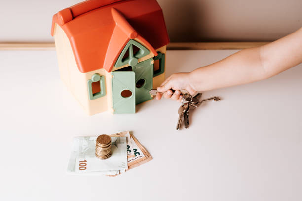 A hand holding keys next to a toy house, with a stack of coins and banknotes on a table.