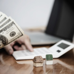 Person holding dollar bills in one hand and using a calculator next to miniature houses.