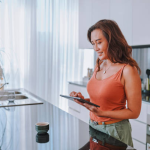 A woman in a modern kitchen reads on a tablet, possibly researching Dubai real estate benefits.