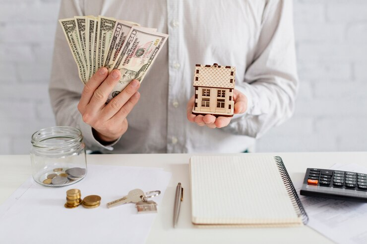 Person holding dollar bills and a small house model, with a jar of coins, keys, a pen, a notebook, and