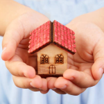 A person is holding a small wooden house with a red roof in their hands.